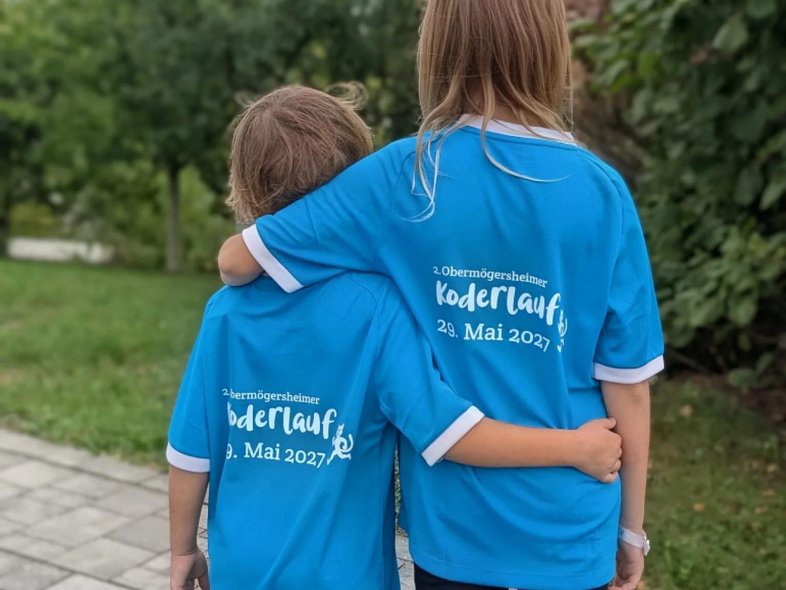 Zwei Kinder von hinten mit hellblauem Koderlauf-Shirt 2027, Rückenmotiv 2. Obermögersheimer Koderlauf 29. Mai 2027
